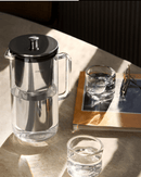 Aarke Purifier carafe with water on a table beside a glass and book, showcasing its sleek glass and stainless steel design.