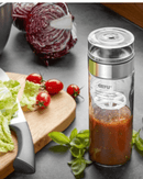 shaker à vinaigrette MIX GEFU en verre 350 ml avec vinaigrette maison et légumes frais en arrière-plan